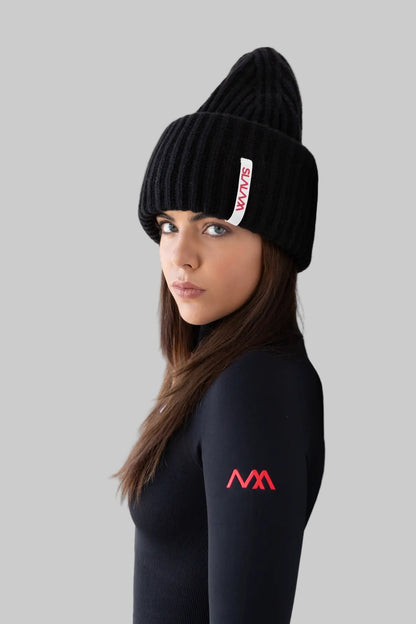 Woman wearing a black beanie and black ski bodysuit with a logo, leaning against a white wall.