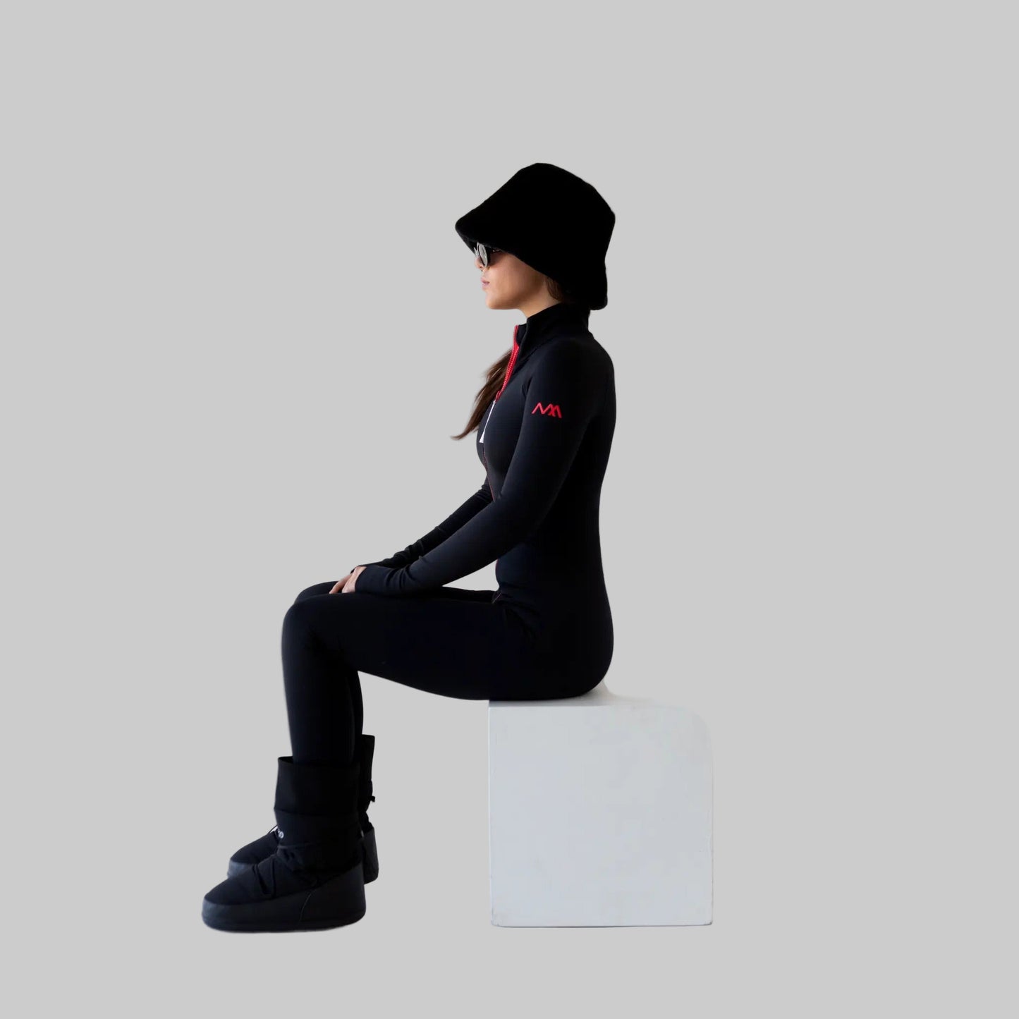 Women wearing a black ski bodysuit with a logo, sitting on a white cube against a white background