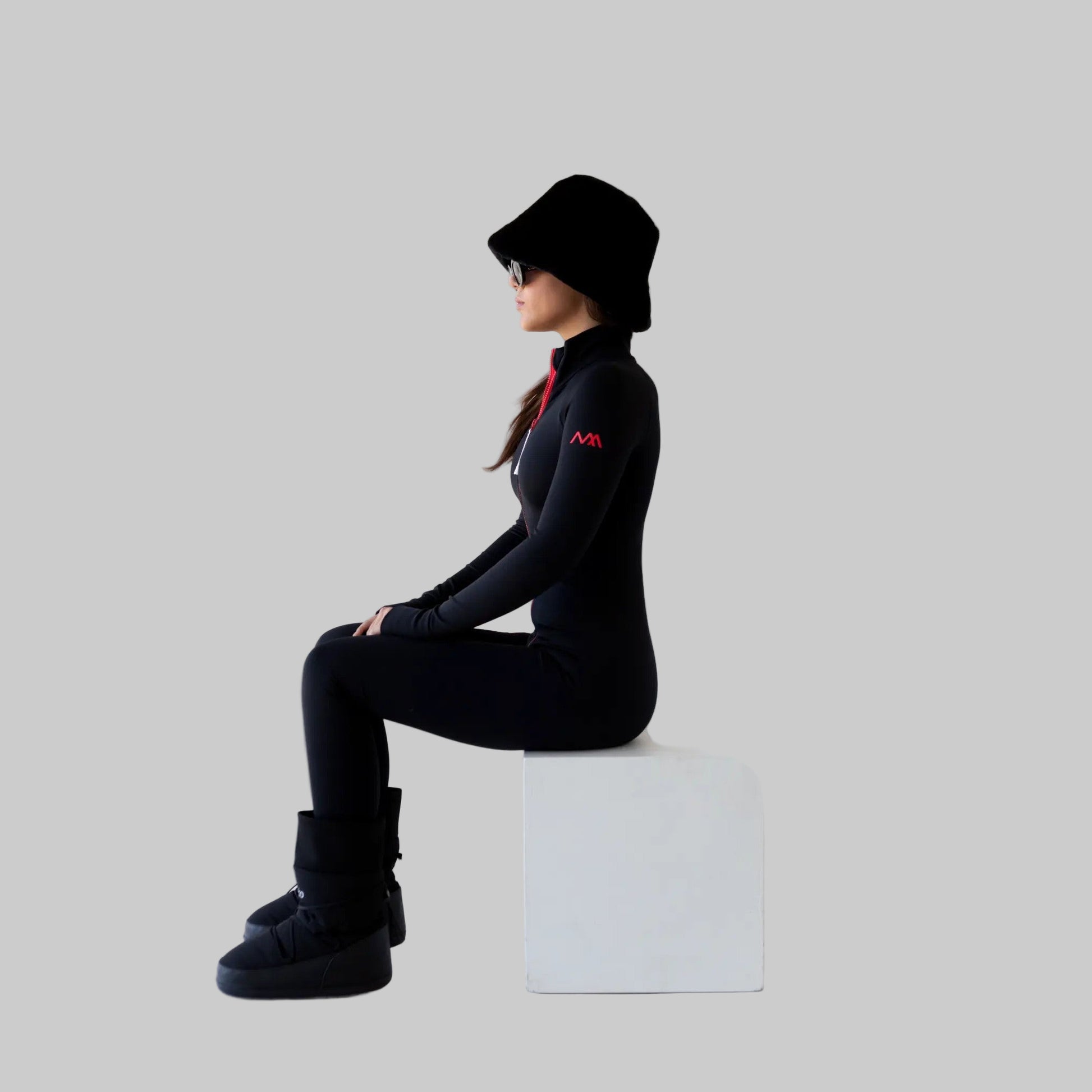 Women wearing a black ski bodysuit with a logo, sitting on a white cube against a white background