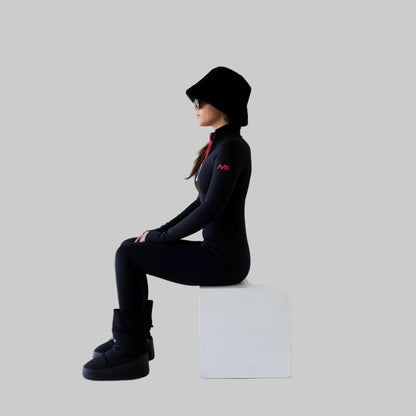 Women wearing a black ski bodysuit with a logo, sitting on a white cube against a white background